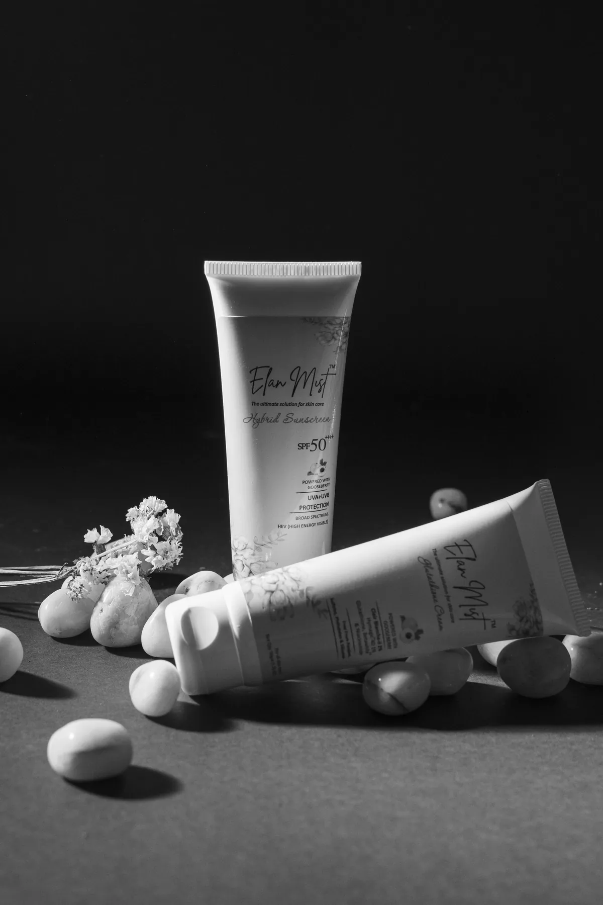 Black-and-white photo of Elanmist Hybrid Sunscreen and Glutathione Cream tubes among pebbles